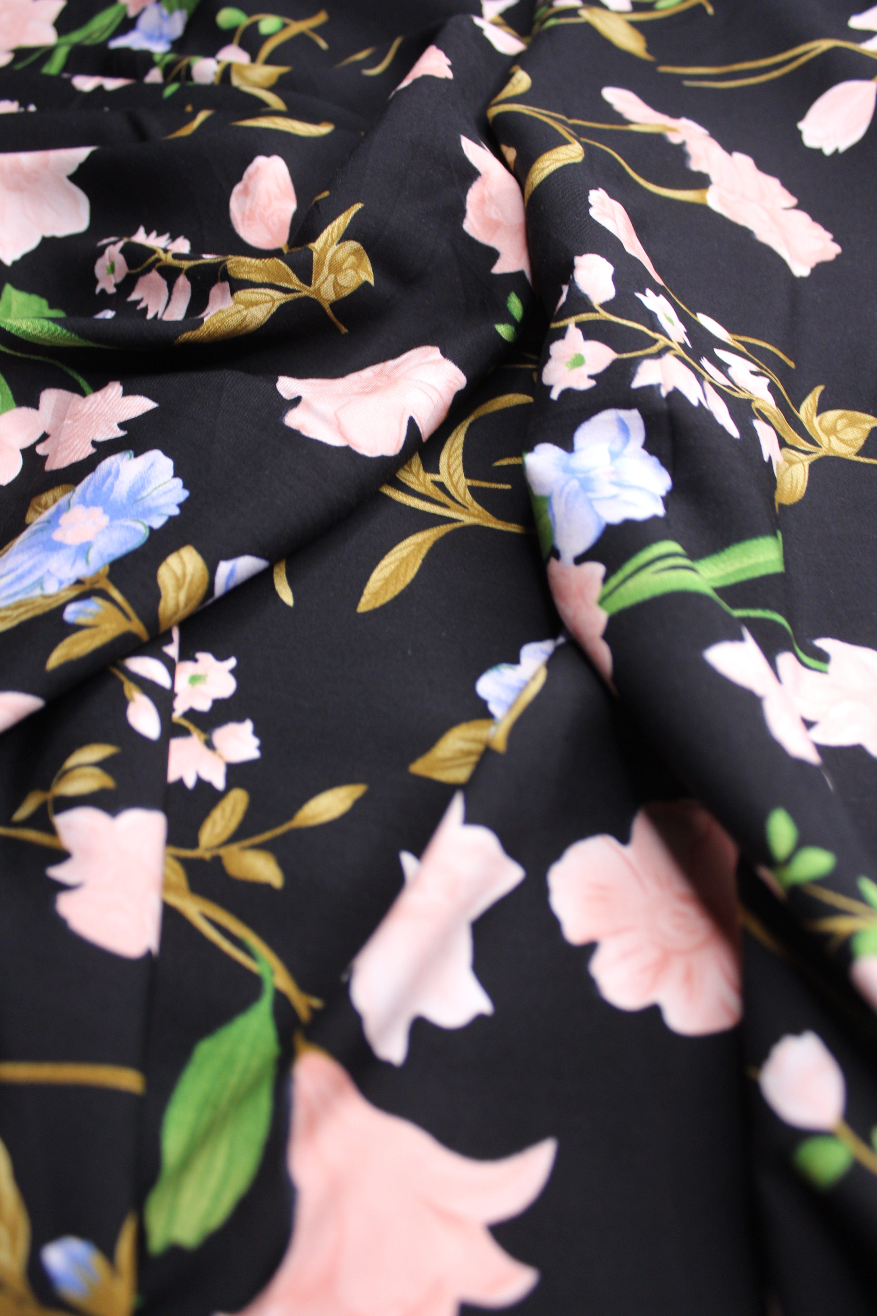 Black fabric with a floral print featuring light pink, white, and blue flowers, green leaves, and gold stems, shown in soft folds.