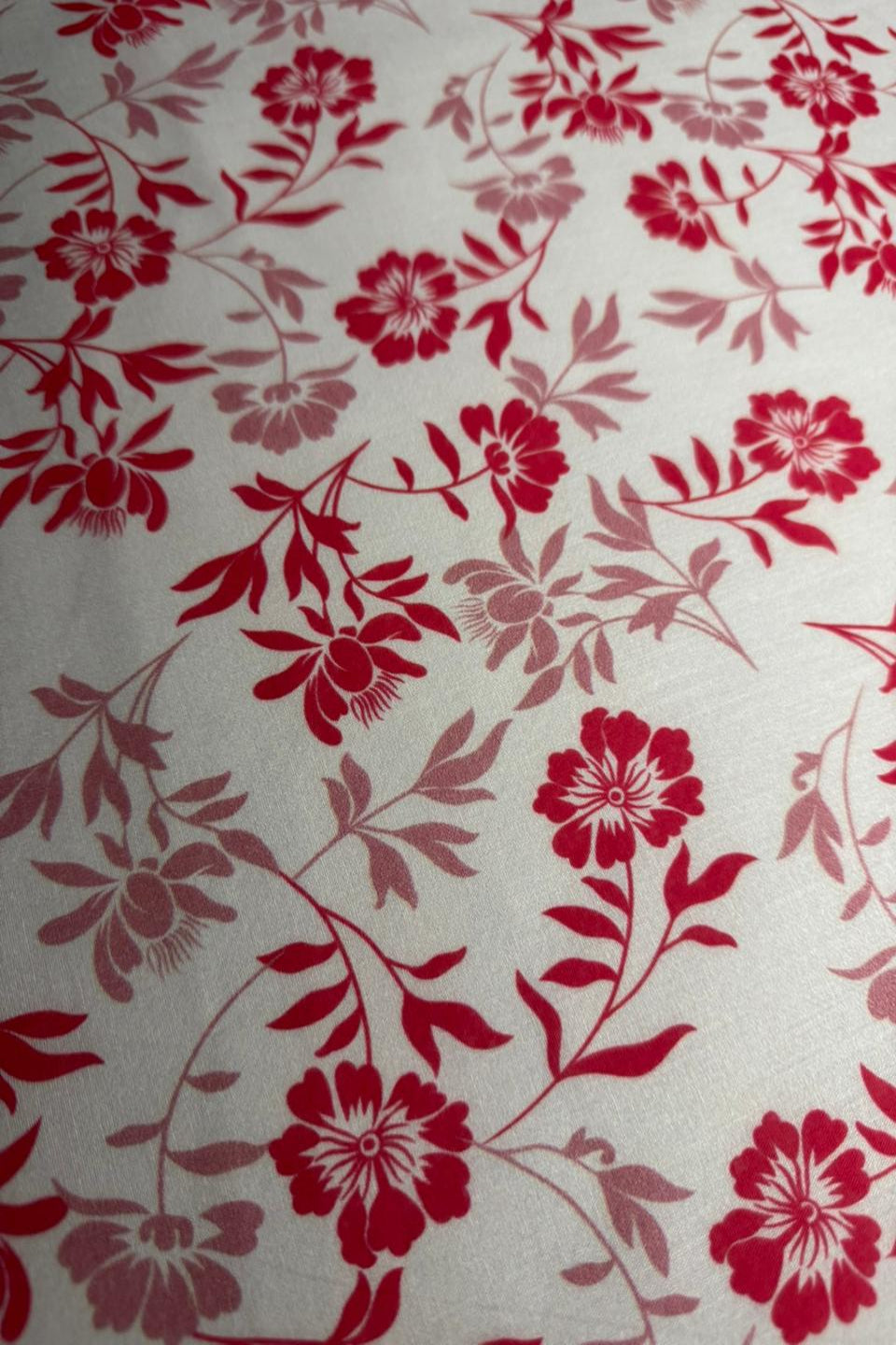 A close-up of a floral patterned fabric or wallpaper featuring red and dusty rose flowers with green leaves on a light cream background.