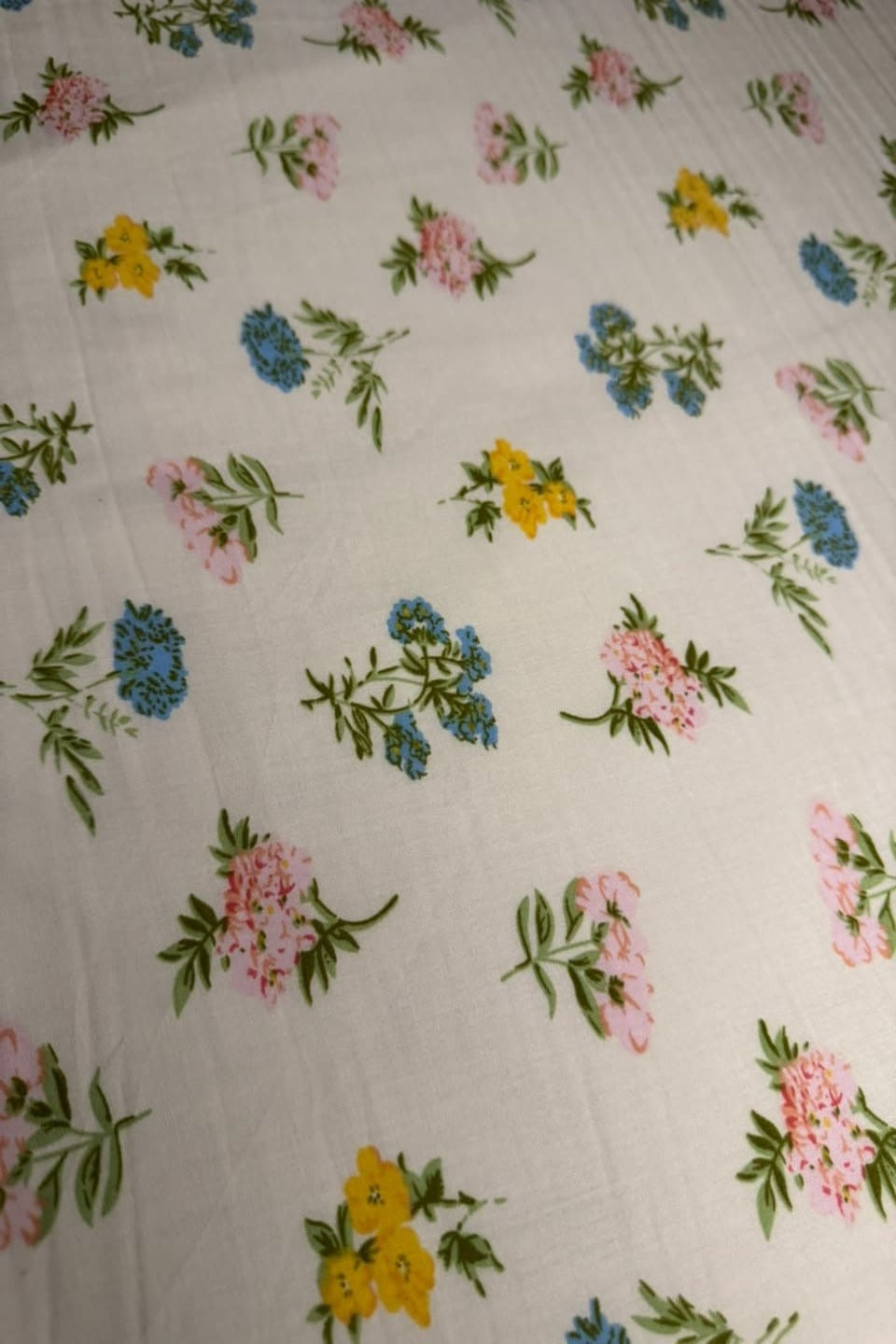 A close-up of a white or off-white fabric with a scattered floral print featuring pink, yellow, and blue flowers with green leaves.
